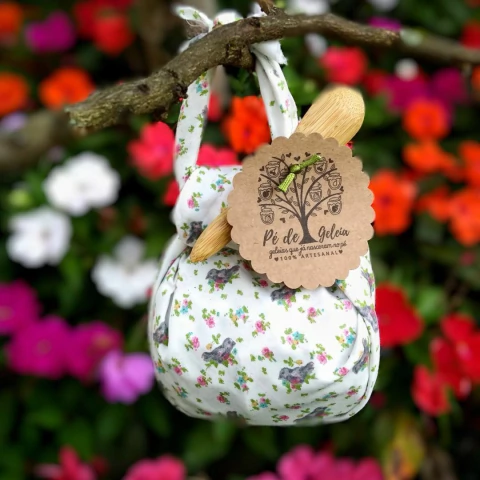 Frasco de geleia embalado em tecido estampado em formato de bolsinha, pendurada em um galho, com flores coloridas ao fundo.