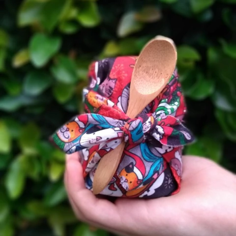 Frasco de geleia envolto em um tecido colorido e estampado, segurado por uma mão, acompanhado de uma colher de bambu.