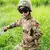 Criança vestindo uniforme de treinamento tático militar, com capacete e óculos de proteção, em meio a grama alta.