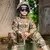 Criança vestindo uniforme de treinamento tático militar, com óculos de proteção e capacete, sentada em um caixa de munição.