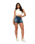 Modelo feminina usando shorts jeans com detalhes desfiados, combinado com um top cropped branco e tênis, posando em um ambiente moderno.