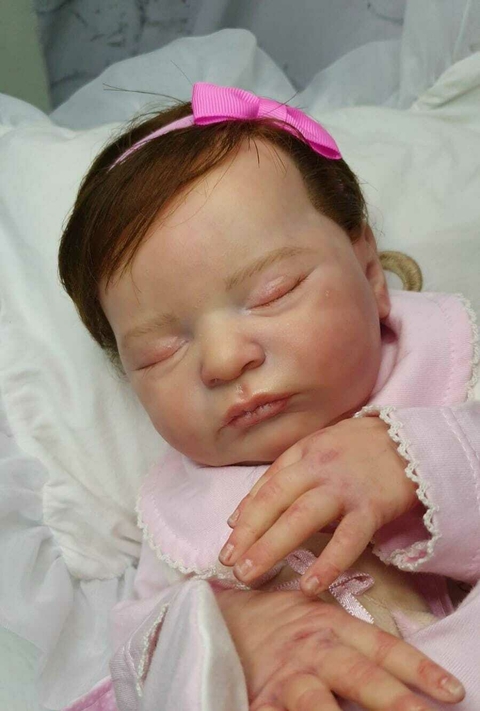 Bebê reborn super realista Laura com cabelo castanho e laço rosa, deitada sobre um tecido branco.