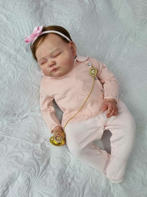 Bebê reborn super realista Bianca vestida com um macacão rosa, usando um laço na cabeça e segurando um chupeta dourada.