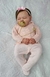 Bebê Reborn super realista Bianca vestindo macacão rosa e chupeta, dormindo em superfície branca com detalhes de bordado.