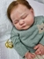 Bebê reborn super realista Brooklyn, com expressão serena, vestindo um suéter verde e segurando um chupeta dourada ao lado.