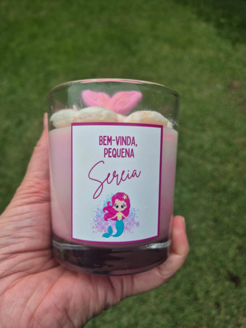 Vela temática artesanal em copo de vidro, com acabamento decorativo em formato de conchas e uma cauda de sereia rosa em destaque. O rótulo personalizado traz a frase "Bem-vinda, pequena sereia" seguida do nome "Maitê", com ilustração de uma pequena sereia