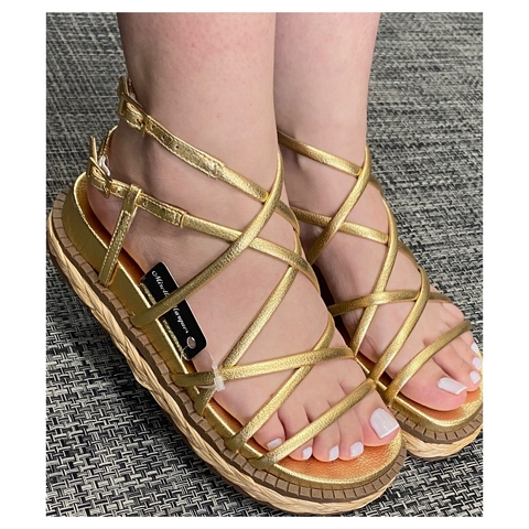 Sandalia Tiras dourada com tiras cruzadas, vista de cima, em um fundo texturizada com detalhe de palha trancada no solado  