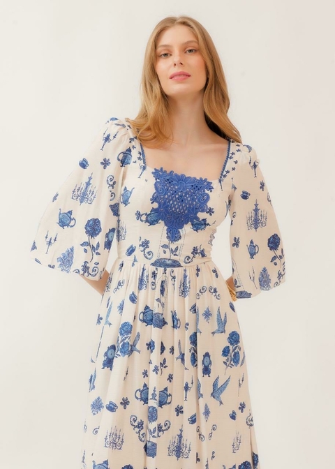 Mulher vestindo vestido midi branco com estampa azul floral e detalhes em renda no busto, com mangas bufantes