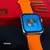 Smartwatch WS25 Mini 42mm com tela de jogos, pulseira laranja e caixa vermelha ao fundo.