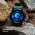 Smartwatch Thunder Wearzone com tela mostrando o menu de controle de músicas, com pulseira verde e corpo preto, sobre pedras.
