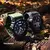 Dois smartwatches Thunder Wearzone com pulseiras robustas, um verde e outro preto, exibidos sobre tronco de árvore em ambiente externo