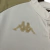 Logotipo dourado da marca Kappa em uma camisa de futebol branca, destacando-se no tecido.