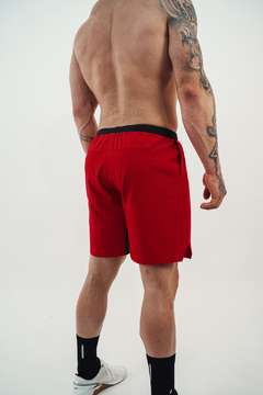 Homem visto de costas vestindo shorts vermelhos com cós preto e tênis branco, em fundo branco
