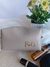 Necessaire box personalizada em material sintético bege com iniciais douradas F e G, exibida sobre superfície clara ao lado de produtos de beleza e vaso com flores