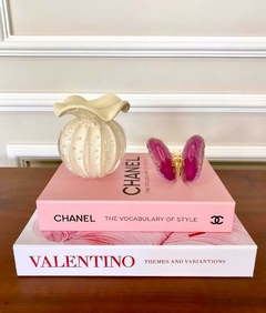 Um vaso de cerâmica marfim com formato de flor, posicionado sobre livros de moda da Chanel e Valentino, em uma mesa de madeira.