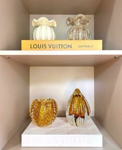 Vasos trouxinha de vidro murano decorativos em diferentes formas e cores, incluindo um vaso marfim e um vaso dourado, exibidos em uma prateleira ao lado de um livro da Louis Vuitton.