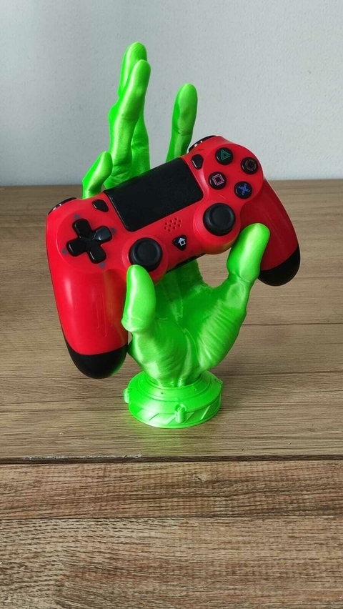 Suporte para joysticks em forma de mão alienígena verde segurando um controle vermelho sobre uma mesa de madeira.