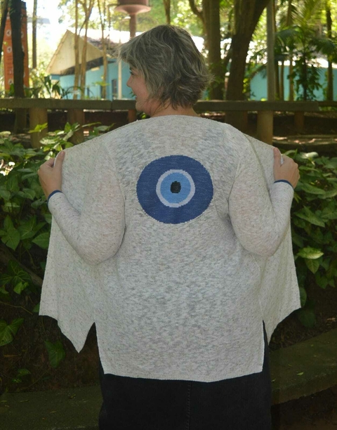 Mulher usando um cardigan de cor clara com um desenho de olho grego nas costas, em um ambiente natural.