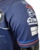 Detalhe da camisa masculina azul da BMW para a temporada de Fórmula 1 23/24, mostrando o logotipo e patrocínios no braço.