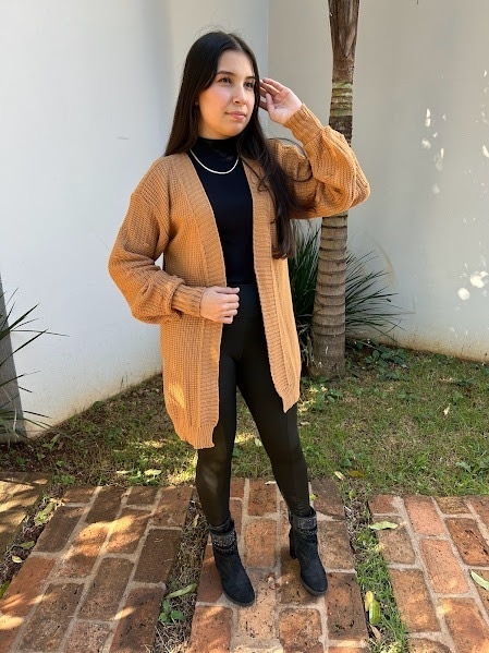 Mulher usando cardigan tricô marrom claro aberto, blusa preta de gola alta, calça preta e botas pretas em jardim com piso de tijolos