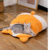 Costalito y camita esponjosa en forma de gatito durmiendo boca abajo, con orejitas, colita y patitas traseras que sobresalen. Con gatito encima.
