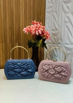 Dupla de bolsas de mão em cores azul e rosa, com detalhes acolchoados e alças douradas, decoradas com um vaso de flores ao fundo.