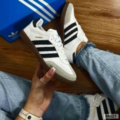 Tênis Adidas Samba OG com detalhes em preto e sola marrom, posicionado em um fundo de madeira.