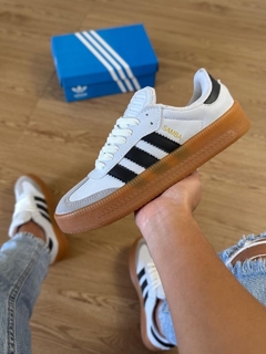 Tênis Adidas Samba OG em destaque, com solado de borracha, parte superior branca e detalhes em preto, sendo segurado por uma mão.