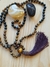 Japamala feito com contas de olho de tigre e ágata, com um pompom roxo, sobre uma superfície de madeira.