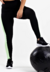 Calça legging preta com listra lateral verde e branca, usada em treino, ao lado de uma bola de exercício.