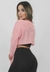 Modelo usando um cropped fitness rosa, com uma blusa de malha que revela a parte inferior da barriga, combinando com calças pretas.