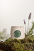 Vela aromática Domus Vitalis em um recipiente de cerâmica com detalhe em verde, cercada por flores e ervas. Inspirada no acorde chipre criado por François Coty