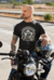 Homem tatuado sentado em uma motocicleta, vestindo uma camiseta preta com o design 'Custom Motors'.