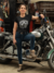 Mulher posando ao lado de uma moto customizada, vestindo uma camiseta preta com a inscrição "Custom Motors".