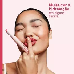 Modelo aplicando o Lip Balm Radiance Balm da Ruby Rose, mostrando a embalagem e um sorriso radiante.