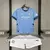 Kit infantil do Manchester City, composto por camisa azul e short branco, exibido em um suporte.