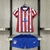 Kit infantil do Atlético de Madrid para a temporada 2024/25, composto por camisa vermelha e branca com detalhes em azul e calção azul.