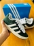 Zapatillas Adidas Campus verde con detalles en blanco, sujetadas por una mano, frente a una caja azul con el logo de Adidas.