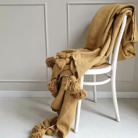 Manta de lanilla color mostaza con pompones, drapeada sobre una silla blanca de madera.