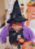Boneca Bruxinha dos Cristais com cabelo roxo, chapéu preto e varinha, acompanhada de itens decorativos como abóbora e gato.