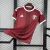Camisa vermelha da seleção do Catar para torcedor, modelo masculino, com detalhes brancos nas mangas e gola, logo da Adidas e emblema da Federação de Futebol do Catar