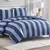 Edredom de casal 100% algodão Doce Vida São Luís MA, com estampa de listras horizontais em dois tons de azul, sobre uma cama. Inclui dois porta-travesseiros coordenados e a cabeceira da cama é branca com detalhe curvo, e um abajur dourado está na mesinha 