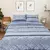 Cama arrumada com edredom Queen Doce Vida São Luís MA e dois porta-travesseiros, apresentando padrões em tons de cinza e azul, incluindo desenhos trançados e geométricos. O ambiente possui decoração minimalista com quadros na parede e plantas.