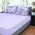 Jogo de lençol King em malha roxo da marca Portallar. O lençol de baixo é na cor lilás liso e as fronhas têm uma estampa de flores roxas sobre um fundo branco. A cama está em um quarto com duas mesas de cabeceira com luminárias e uma janela ao fundo. Envi