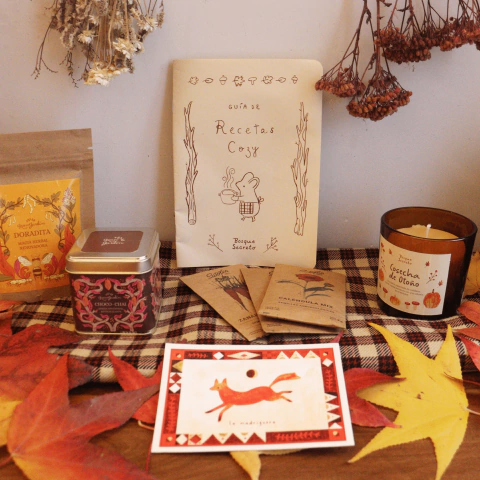 Cajita de productos cozy "Momentos en la Madriguera" con infusiones, vela aromática, semillas para plantar y guía de recetas sobre un mantel a cuadros con hojas secas otoñales