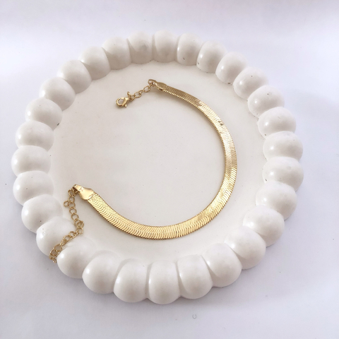 Pulseira de fita laminada em ouro 18K com brilho, disposta em um suporte branco.