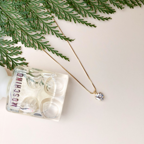 Colar ponto de luz em forma de coração em ouro 18K com zircônia, posicionado ao lado de um frasco de perfume Moschino e folhas verdes.