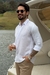 Homem sorrindo em um barco, vestindo uma camisa de linho branca e calças bege, segurando uma taça de champanhe.