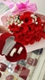 Par de alianças de namoro grossas fosqueadas 8mm dentro de estojo vermelho em formato de coração, ao fundo um buquê de rosas vermelhas com detalhes em branco e fita rosa