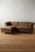 Sillón esquinero Chesterfield en tono marrón, con diseño clásico, tapizado en ecocuero y detalles de acolchado, situado sobre un suelo de madera clara.
